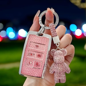 May include: A pink car key fob cover with a sparkly finish, silver frame and buttons. A matching pink crystal bear keychain hangs from the fob. The key fob has the text "RADIANT KEY" in the upper left corner.