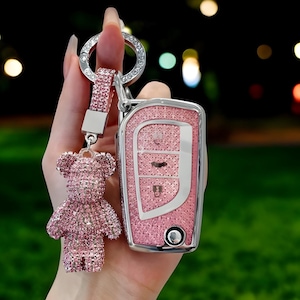 Op de afbeelding: Roze glinsterende autosleutelketting met een roze glinsterende teddybeerbedel. De sleutelketting heeft een zilveren ring en een zilveren band. De autosleutel is ook roze en glinsterend.