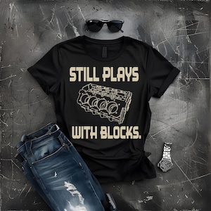 May include: Black t-shirt with the text "STILL PLAYS WITH BLOCKS" and a diagram of an engine block. The image also includes a pair of blue jeans, sunglasses, and a wristwatch.