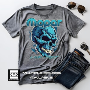 May include: Gray t-shirt with a blue skull graphic and the text "Mopar Live Fast". The shirt has a distressed look and the text "Multiple Colors Available" is printed below the graphic.
