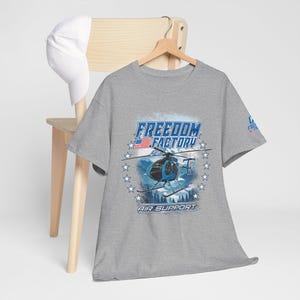 May include: A heather gray short-sleeve t-shirt featuring a blue helicopter graphic, the text Freedom Factory Air Support, and a small American flag. A white baseball cap hangs on the wooden hanger behind the shirt, which displays the logo.