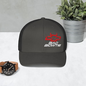 May include: A charcoal gray trucker hat with a black mesh back. The front of the hat features the text "BAD BOWTIE" in white, with a red graphic above it. A wristwatch with a brown leather band is in the foreground.