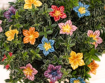 May include: A variety of colorful ceramic flowers are arranged in a green bush. The flowers are in shades of pink, blue, yellow, orange, and red.