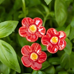 May include: Three red ceramic flower-shaped garden markers with yellow centers. The markers are arranged in a cluster and are surrounded by green foliage.