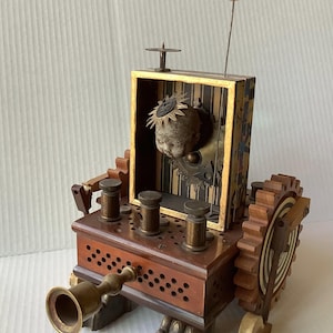 May include: A mixed-media sculpture featuring a doll head inside a rectangular box with a striped interior. The base is a dark wood block with three brass cylinders, a brass horn, and a large gear on the side. It stands on four claw-shaped feet.