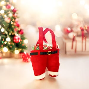 Op de afbeelding: Rode kerstmanbroekjes-cadeauzak met witte bies en een zwarte riem. De tas is gevuld met kleurrijk tissuepapier en klaar voor cadeaus.
