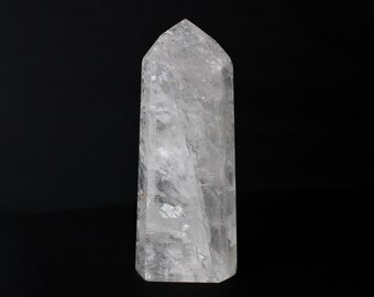 May include: A clear quartz crystal point with a faceted top. The crystal is transparent with internal inclusions and sits against a black background. This crystal is a decorative item.