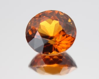 May include: A faceted, oval-cut gemstone with a warm, amber-orange hue. The gem's multiple facets reflect light, creating a brilliant sparkle. The gem is displayed on a reflective surface, showing its mirrored image.