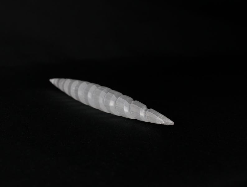 May include: A white, segmented, tapered object, possibly a sculpture or decorative item, against a black background. The object has a series of rounded, ridged segments that narrow to a point. The lighting creates highlights and shadows, emphasizing the texture.