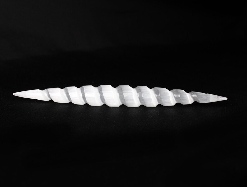 May include: A white, spiraled, selenite crystal wand. The wand has a pointed tip on both ends and a ridged, twisted design along its length. The background is solid black.