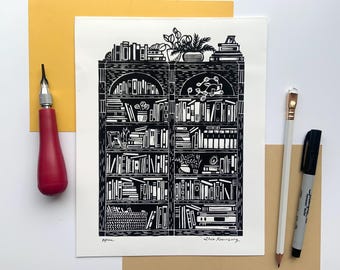 May include: Black and white linocut print of a bookshelf filled with books and plants. A red carving tool, a white pencil, and a black marker are on the table. The print is signed by the artist.