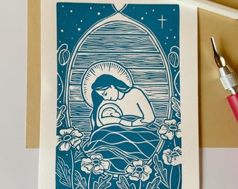 May include: A blue and white linocut print of a woman holding a baby. The woman is surrounded by flowers and the print is titled "Comfort + Joy" and signed "Shen Pearbury".