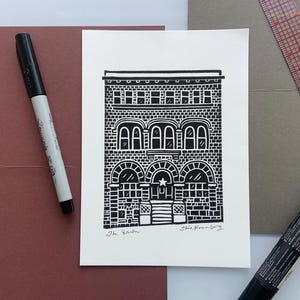 May include: A black and white ink drawing of a building with arched windows and a star above the entrance. The artwork is on white paper, with a black marker and other stationery items in the background.