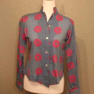 May include: A button-down shirt with a mandarin collar, displayed on a mannequin. The shirt is a light blue with a pattern of large, bright pink circles. The long sleeves have pink trim at the cuffs.
