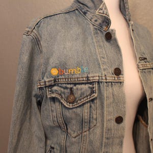 May include: A light blue denim jacket with a colorful embroidered patch that says "bumble" on the left chest pocket.