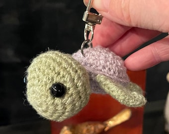 May include: A crocheted turtle keychain with a purple shell and green head and feet. The turtle has black eyes and is attached to a silver key ring.