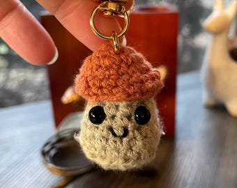 May include: A crocheted mushroom keychain with a smiling face and black eyes. The mushroom has a brown cap and a beige body. The keychain is attached to a gold metal ring.