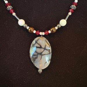 Può includere: Una collana con un pendente ovale con un motivo bianco e grigio. La collana ha perline rosse, bianche e dorate, con dettagli in metallo argentato. Il pendente è sospeso da un anello di metallo argentato.