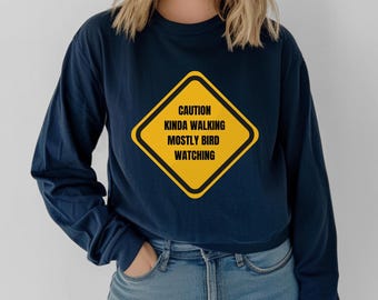 May include: Navy blue long-sleeved shirt with a yellow caution sign graphic that reads "Caution Kinda Walking Mostly Bird Watching".