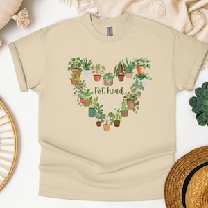 May include: Beige t-shirt featuring a heart-shaped arrangement of potted plants. The words "Pot head" are printed in green within the heart. The shirt is made of soft material and has a classic crew neck.