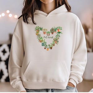 May include: Cream-colored hoodie with a front pocket and a hood. The design features a heart-shaped arrangement of potted plants, with the words "Pot head" in the center. The plants are in various pots and include different types of greenery.