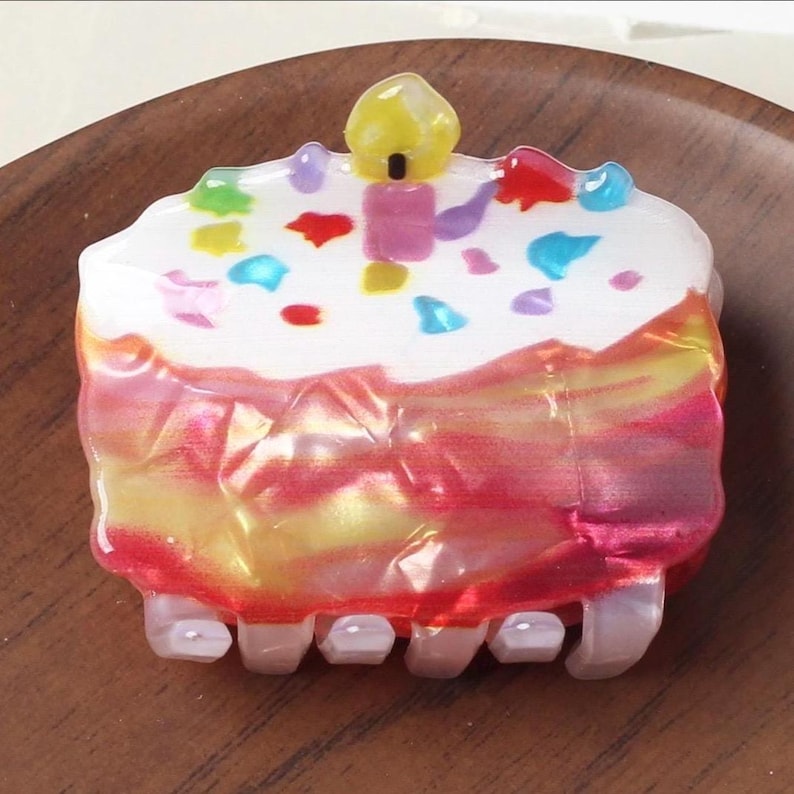 May include: A cake-shaped hair clip with a white frosted top, colorful sprinkles, and a yellow candle. The clip has a translucent, layered design with red, pink, and yellow hues. The clip is approximately 1.5 inches wide.