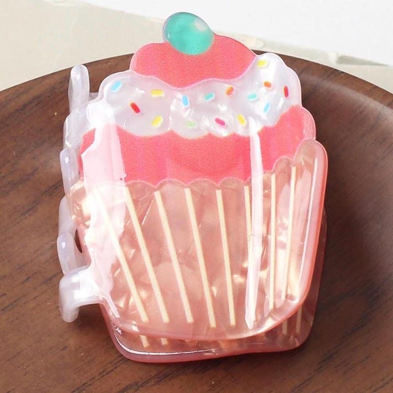 May include: A cupcake-shaped hair clip with a pink frosted top, a green cherry, and colorful sprinkles. The clip has a clear, pink, and white design with vertical lines on the base, resembling a cupcake liner. The clip is made of plastic.