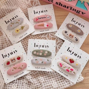 May include: A selection of handmade hair clips with embroidered floral and fruit designs. The clips are in shades of pink, green, and beige, and the packaging features the word "bunny". The designs include flowers, strawberries, and a bear.