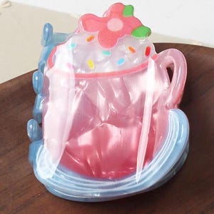May include: A pink and blue hair clip shaped like a cup of coffee or tea, topped with a pink flower and sprinkles. The clip has a handle and sits on a blue saucer. The design is whimsical and colorful.