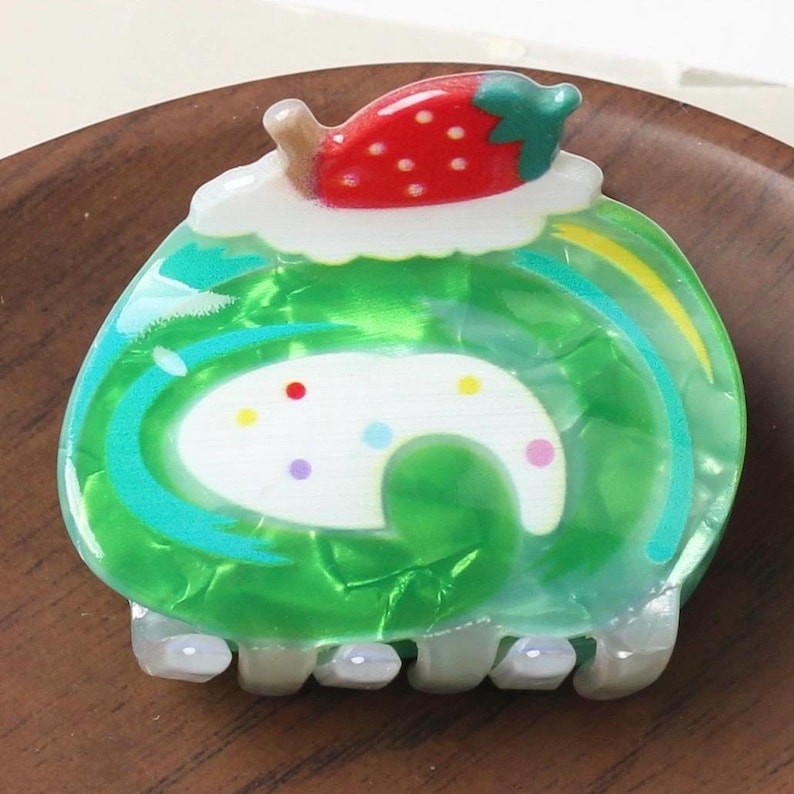 May include: A green, swirl-patterned hair clip shaped like a cake roll, topped with a red strawberry and white frosting. The clip has a white base with colorful polka dots and a claw-style grip.
