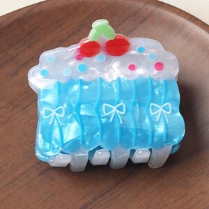 May include: A decorative hair clip in the shape of a cupcake. The clip features a blue, ruffled design with white bows, topped with a clear frosting layer and red cherry accents. The clip is made of plastic.