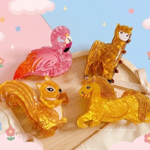 May include: Four animal-shaped hair clips: a pink flamingo, a golden squirrel, a golden llama, and a golden horse. The clips are made of a translucent material and are arranged on a wooden tray with a ribbon.