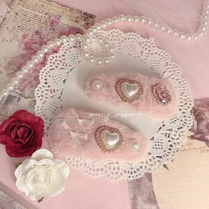 Può includere: Due fermagli per capelli rosa e pelosi con decorazioni di perle e strass. I fermagli sono su un pizzo bianco con una collana di perle e uno sfondo rosa.