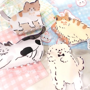 May include: Four decorative hair clips in the shape of cartoon animals. The clips include a calico cat, a tabby cat, a black and white cat, and a white dog with a bow tie. The background includes pastel colored patterned fabric.