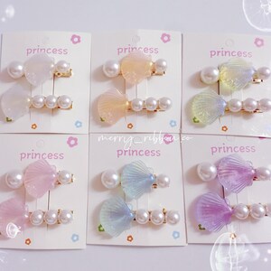 Puede incluir: Seis juegos de pinzas para el pelo con temática de princesa. Cada juego incluye una pinza en forma de concha y una pinza en forma de lazo, ambas adornadas con perlas de imitación. Las pinzas vienen en varios colores pastel, incluyendo rosa, amarillo y azul. La palabra "princess" está impresa en las tarjetas.