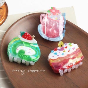 May include: Three colorful acrylic hair clips shaped like desserts: a green swiss roll with a strawberry, a pink drink with a flower, and a slice of cake with sprinkles. The text "merry_ribbon.co" is visible on the wooden tray.