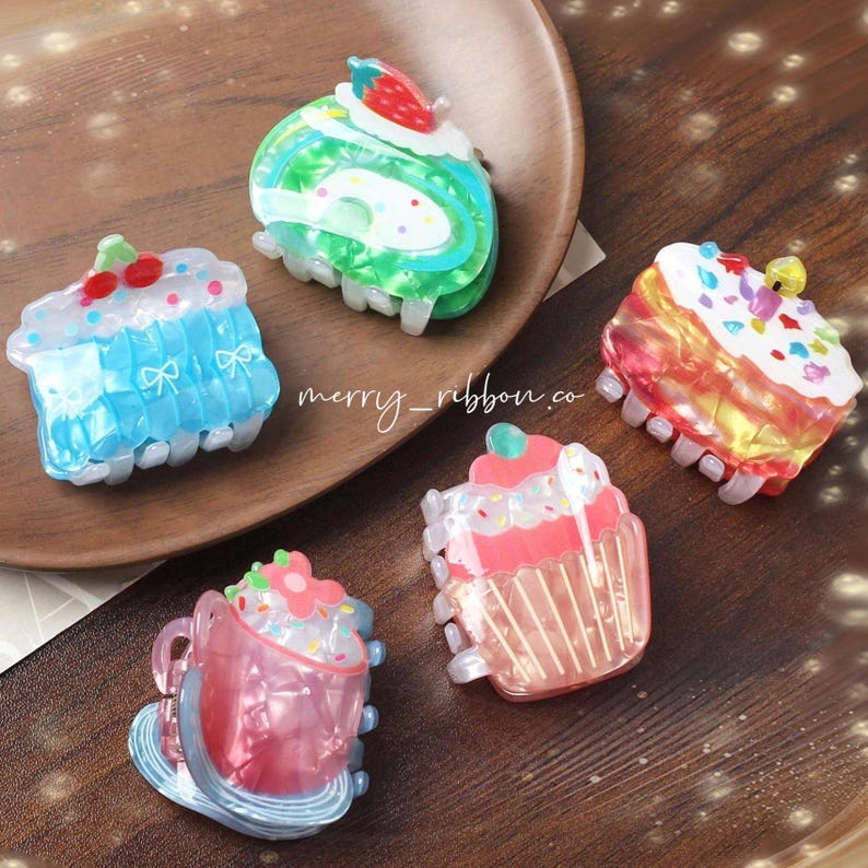 May include: A collection of colorful, dessert-themed hair clips. The clips are shaped like cupcakes, cakes, and a cup of tea, with details like sprinkles, bows, and strawberries. The text "merry_ribbon.co" is visible.