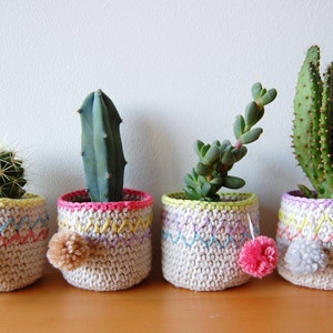 May include: Four small, handmade crochet plant pots with cacti and succulents. The pots are cream-colored with colorful embroidered details and pom-poms. Each pot has a different plant, including a round cactus, a tall cactus, and a succulent.