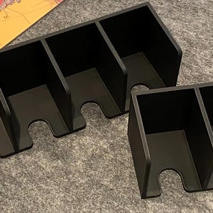 May include: Black organizer trays with multiple compartments. One tray has four sections, and the other has two. Each section has a curved cutout at the bottom. The trays are designed to hold game pieces or other small items.