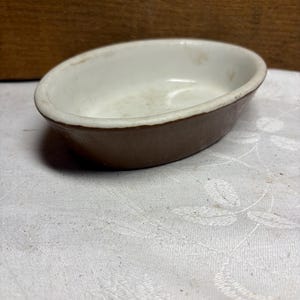 May include: An oval-shaped ceramic dish featuring a smooth, dark brown exterior and a contrasting white interior. The bowl has a simple, rounded rim and a shallow basin, presenting a clean, functional design with a visible color separation.