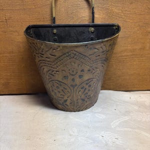 May include: A wall-mounted metal planter with a semi-circular opening and a curved handle. The exterior features a dark, embossed floral pattern on a brass-toned surface, with visible signs of wear and dark patina throughout the metal finish.