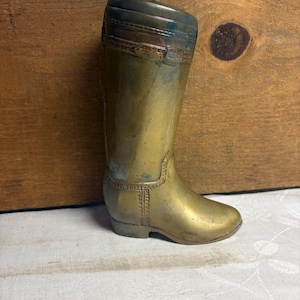 May include: A brass boot-shaped container with a removable lid featuring a small knob handle. The metal surface displays a weathered patina with visible engraved details including a decorative strap and buckle design around the upper shaft.