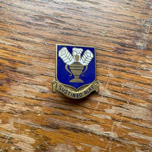 May include: A rectangular pin with a gold-colored border. The pin features a blue shield with a gold-colored vase and three white floral designs. The words "SUSTINEO ALAS" are inscribed on a gold banner below the shield.