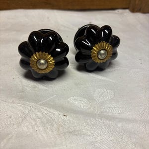 Pode incluir: Dois puxadores de gaveta pretos com acabamento brilhante e formato de flor. Cada puxador apresenta um detalhe central em metal dourado com um pequeno círculo prateado no meio, proporcionando um contraste decorativo à superfície.