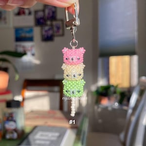 May include: A beaded keychain featuring three stacked bear faces in pink, yellow, and green. The bears have black bead eyes and are attached to a silver-colored clasp. The text "nyasthebeads" and "#1" are visible.