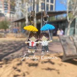 May include: Two beaded figures holding umbrellas, one with a yellow umbrella and the other with a blue umbrella. The figures are wearing school uniforms and are holding hands. The text "Lovely Runner" is below the figures.