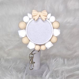 Op de afbeelding: Een sleutelhanger van witte en beige siliconen kralen met een witte cirkel in het midden en een beige strik bovenop. De sleutelhanger heeft een zilveren sleutelring.