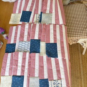 K&ouml;nnte beinhalten: Ein Patchwork-Quilt in Arbeit, mit rot-wei&szlig; gestreiftem Stoff, rot-wei&szlig;em Vichy und blauen Stoffflicken mit wei&szlig;en Punkten und floralen Mustern. Der Quilt liegt auf einer Holzoberfl&auml;che.