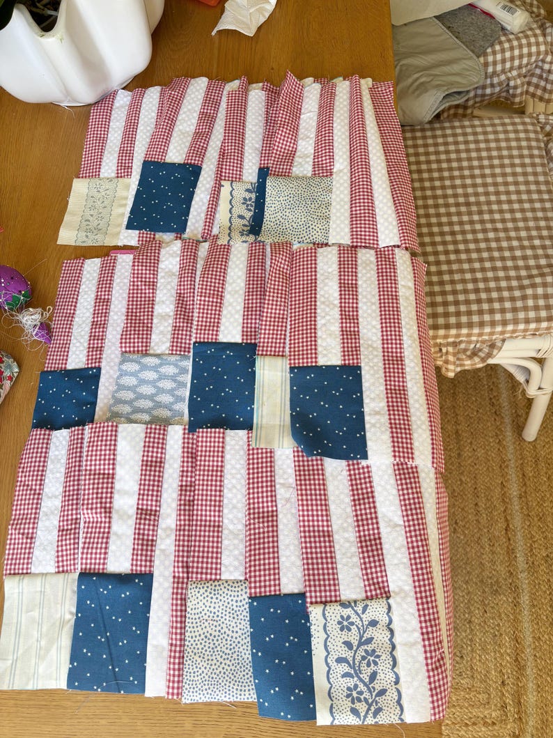 K&ouml;nnte beinhalten: Ein Patchwork-Quilt in Arbeit, mit rot-wei&szlig; gestreiftem Stoff, rot-wei&szlig;em Vichy und blauen Stoffflicken mit wei&szlig;en Punkten und floralen Mustern. Der Quilt liegt auf einer Holzoberfl&auml;che.