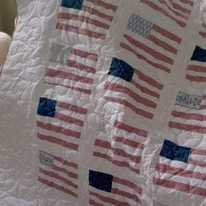 K&ouml;nnte beinhalten: Wei&szlig; gesteppte Decke mit mehreren amerikanischen Flaggenmotiven. Die Flaggen haben rot-wei&szlig; gestreifte Muster mit blauen quadratischen Feldern. Die Decke hat einen rot-wei&szlig; karierten Rand und ein Steppmuster.